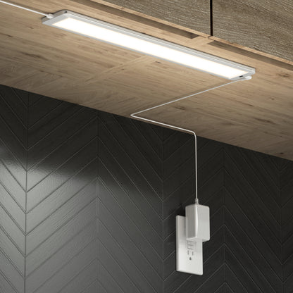 An Artika Maestro under cabinet LED light 3CCT is installed beneath a wooden cabinet, with its power cord plugged into a white outlet on a black chevron-patterned wall.