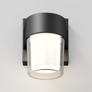 The Artika Halden indoor-outdoor LED wall light features a matte black base and a cylindrical frosted glass shade, providing a soft, warm glow against a white wall.