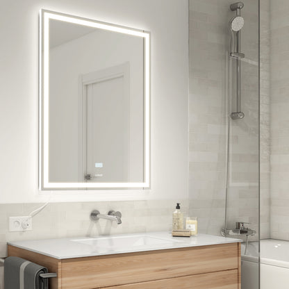 A modern bathroom features a light wood vanity, white sink, wall-mounted faucet, and the Artika Aurea anti fog LED wall mirror. The shower includes a rainfall showerhead and light gray subway tiles. Soap and lotion rest on the counter.