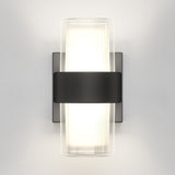 A modern wall sconce with a frosted cylindrical glass shade, mounted on a rectangular metal backplate, featuring a black horizontal band securing the glass. The light glows softly against a white wall.