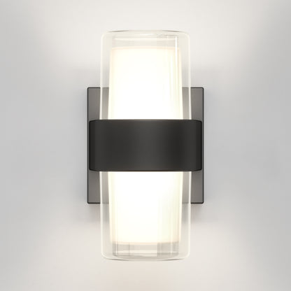 The Artika Colton modern LED indoor-outdoor wall light 5 CCT features a frosted cylindrical glass shade with a black center band and rectangular base, mounted on a light gray wall, casting a soft white glow.