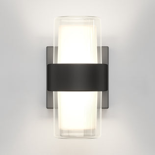 The Artika Colton modern LED indoor-outdoor wall light 5 CCT features a frosted glass cylinder illuminated and held by a wide matte black band, mounted on a rectangular backplate against a light gray wall.