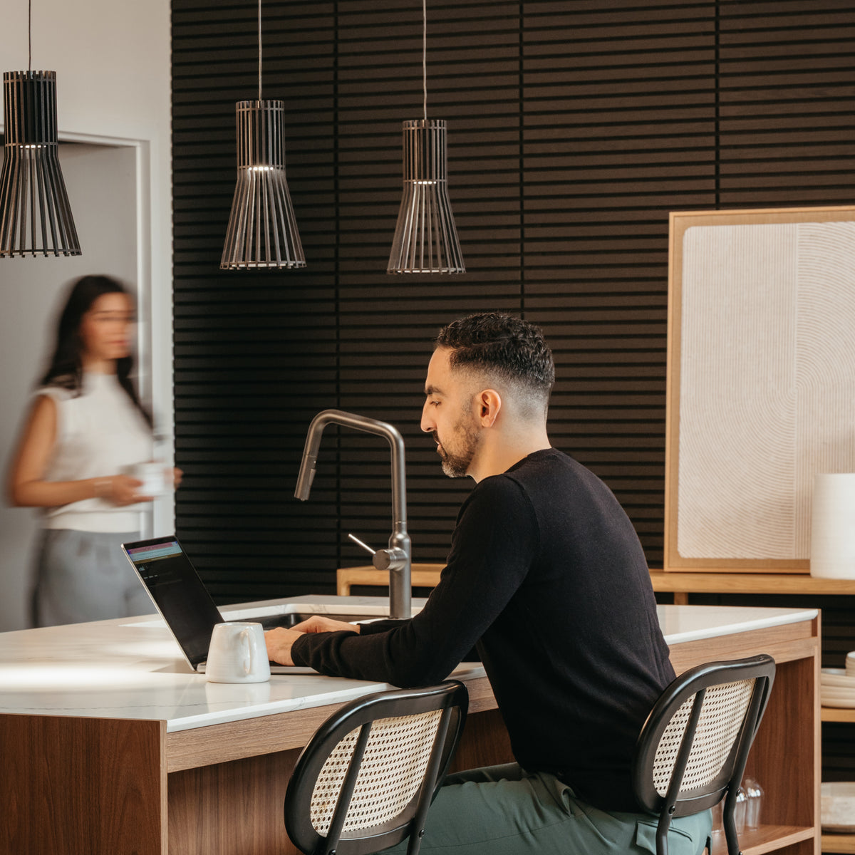 A man sits at a kitchen island working on a laptop, with a coffee mug beside him. Pendant lights hang overhead. In the background, a woman walks past, holding a cup. The space has modern décor with wooden accents.