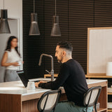 A man sits at a kitchen island working on a laptop, with a coffee mug beside him. Pendant lights hang overhead. In the background, a woman walks past, holding a cup. The space has modern décor with wooden accents.