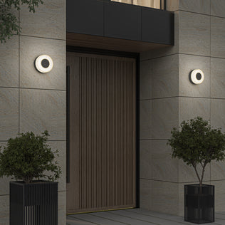 A large wooden door is framed by two Artika Ludwig modern LED indoor-outdoor wall lights (5CCT), with black potted plants set in front of the stone-tiled wall.