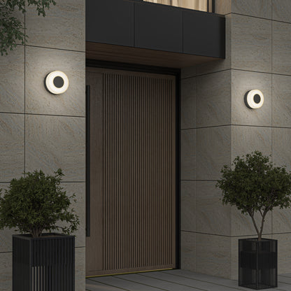 A large wooden door is framed by two Artika Ludwig modern LED indoor-outdoor wall lights (5CCT), with black potted plants set in front of the stone-tiled wall.