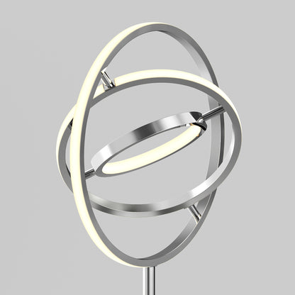 The Artika Pivot modern LED floor lamp 3 CCT features three intersecting metallic rings, two with LED lights, giving it a sculptural and futuristic look against a simple gray backdrop.