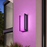 A modern black wall sconce with a rectangular design glows with soft pink light, mounted on a textured, light-colored exterior wall near large windows and green foliage.