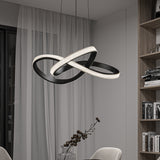 A modern dining room features a sculptural black LED chandelier with intertwined loops, hanging above a table with a glass vase and decorative branches. Sheer curtains and shelves with books are visible in the background.