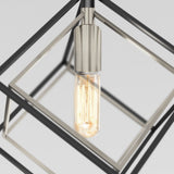 A lit Edison-style light bulb hangs inside a modern geometric metal frame, featuring intersecting square shapes in silver and black against a plain light background.