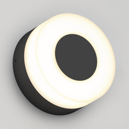 The Artika Ludwig modern LED indoor-outdoor wall light 5CCT features a round black central circle and outer ring, emitting a soft, even white glow, and is shown mounted on a light gray wall.