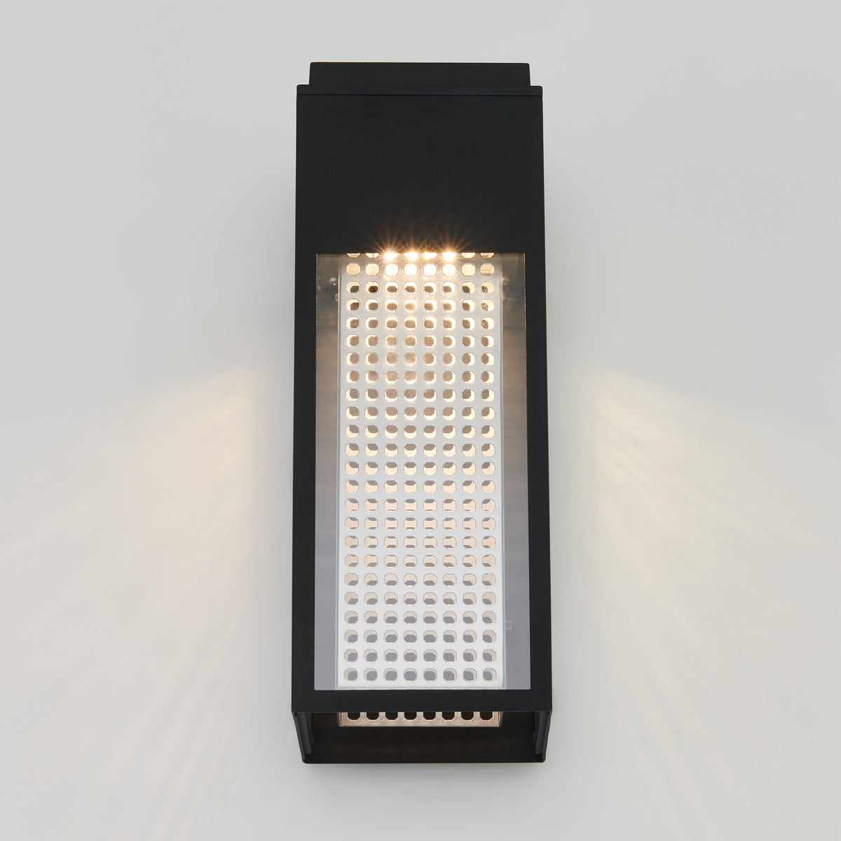 A modern rectangular wall sconce with a black frame and a perforated metallic panel, emitting light from the top and casting subtle patterns on a plain white wall.