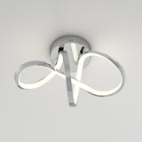 A modern, ceiling-mounted LED light fixture with sleek, chrome curved arms forming an abstract, looped design, radiating a soft white glow against a plain light-colored ceiling.