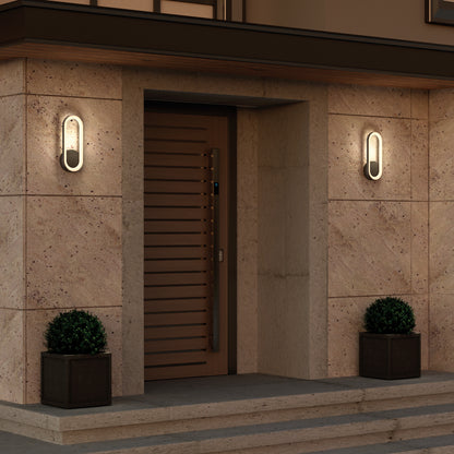 Modern front entrance with a wooden door, stone walls, steps, and two Artika Aiden modern LED indoor-outdoor wall lights on either side. Black square planters with potted plants flank the doorway.