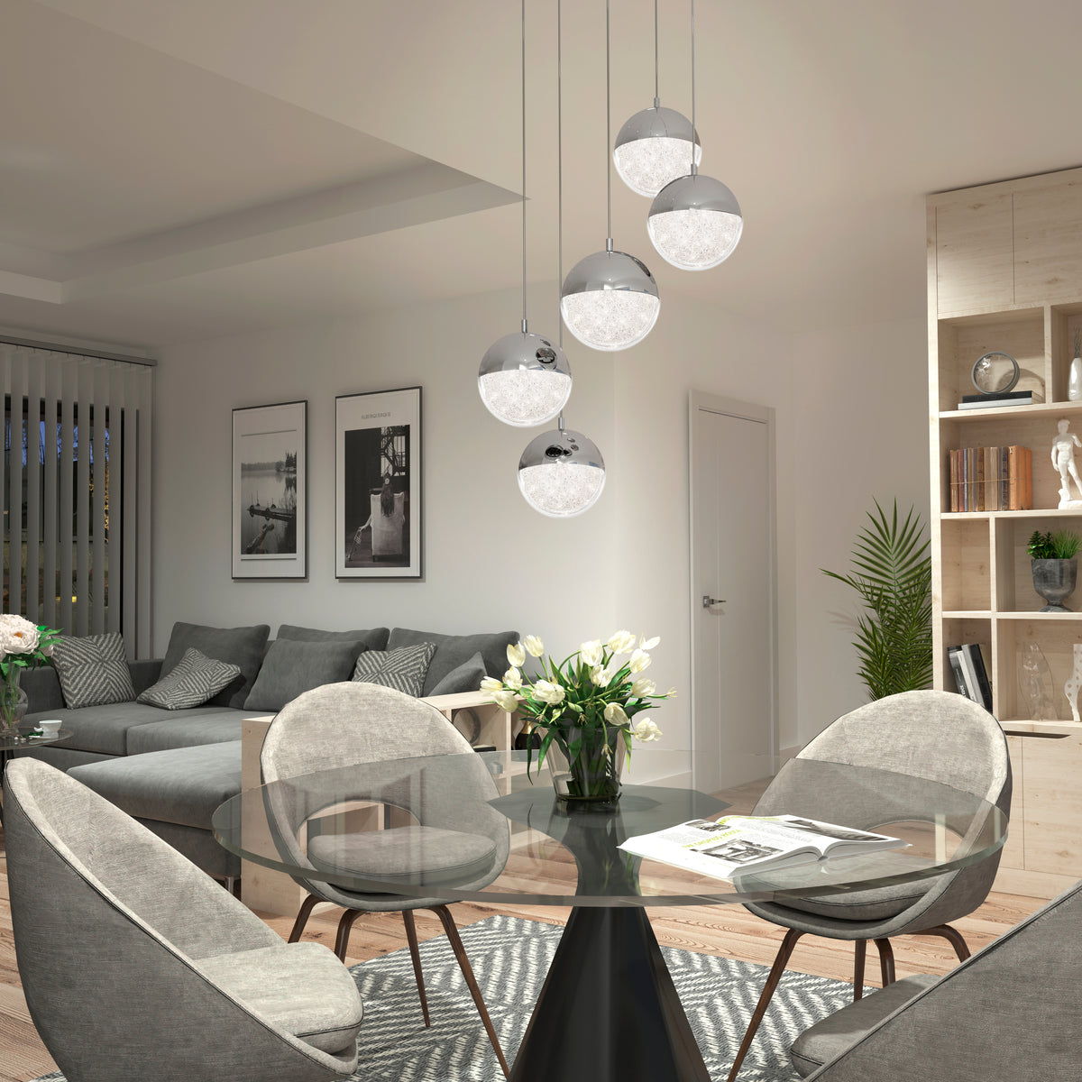 A modern dining and living area with a glass table, four gray chairs, a gray sofa, pendant globe lights, wall art, indoor plants, and wooden shelving with books and decor.