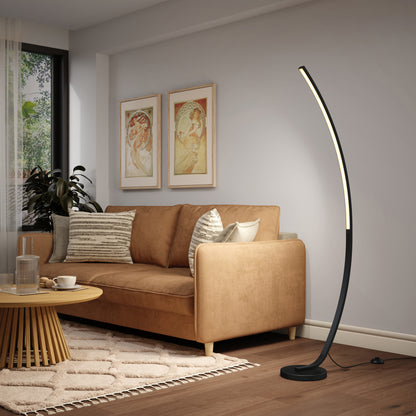 A modern living room features a tan sofa, beige and patterned pillows, a round wooden coffee table, two framed art prints, and the Artika Arc LED Floor Lamp with an arched design standing tall beside the couch.