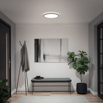 A modern entryway with the Artika Macy 15 in. LED modern flush mount light RGB Wifi, a coat rack with a gray jacket, monochrome abstract art, a bench, small bowl, and potted plant on the floor.