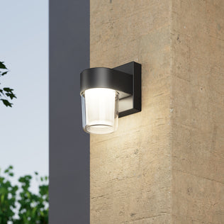 The Artika Halden indoor-outdoor LED wall light 5 CCT in black and clear is mounted on a textured beige wall, glowing at dusk with greenery and a blue sky visible in the background.