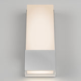 A modern wall sconce with a rectangular frosted glass shade and a brushed metal base, emitting a soft, warm light against a plain light gray wall.