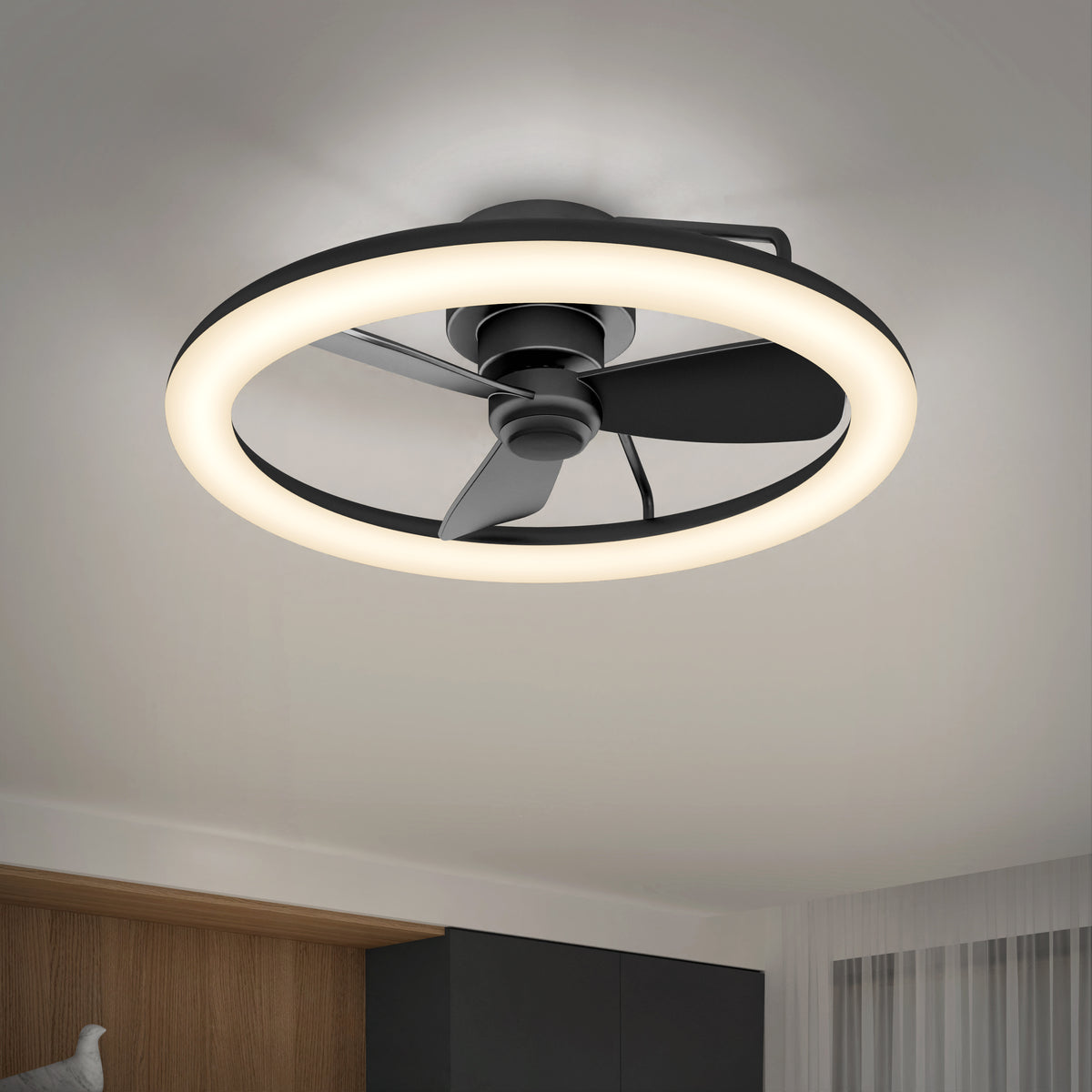 A modern ceiling fan with three black blades is encircled by a glowing LED ring light, mounted on a white ceiling in a contemporary room with minimal decor.