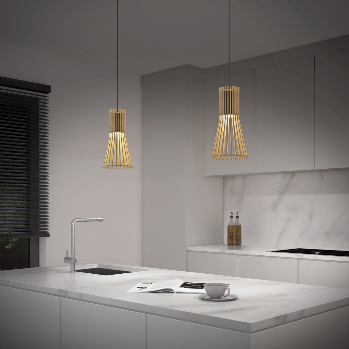 Minimalist kitchen with white cabinets and marble backsplash, featuring two modern pendant lights over a white island. A cup, saucer, and open magazine sit on the countertop. The room is softly lit.