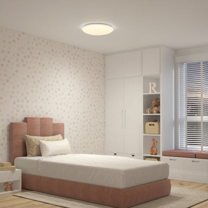 A cozy bedroom with a single bed, pink upholstered headboard, white bedding, and built-in white cabinets. The Artika Empress 13 in. LED modern flush mount light brightens the space with adjustable ambiance. A window seat lets in daylight.