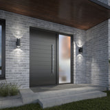 Modern front entrance with a sleek black door, a large frosted glass sidelight, gray brick exterior walls, two wall-mounted lights, and a small porch with steps, surrounded by grass and minimal landscaping.