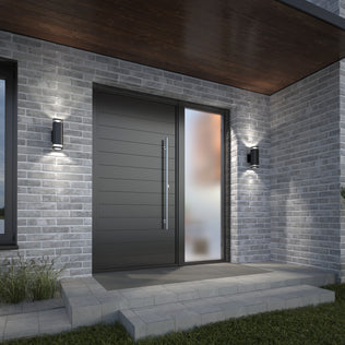 Modern front entrance with a large dark door, vertical handle, frosted glass side panel, gray brick walls, concrete step with grass at the side, and two Artika Mettle LED indoor-outdoor wall lights.