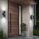 Modern house entrance with a large wooden door, vertical glass panel, two black wall lights, a potted plant on the porch, and light-colored stone walls surrounded by green shrubs.