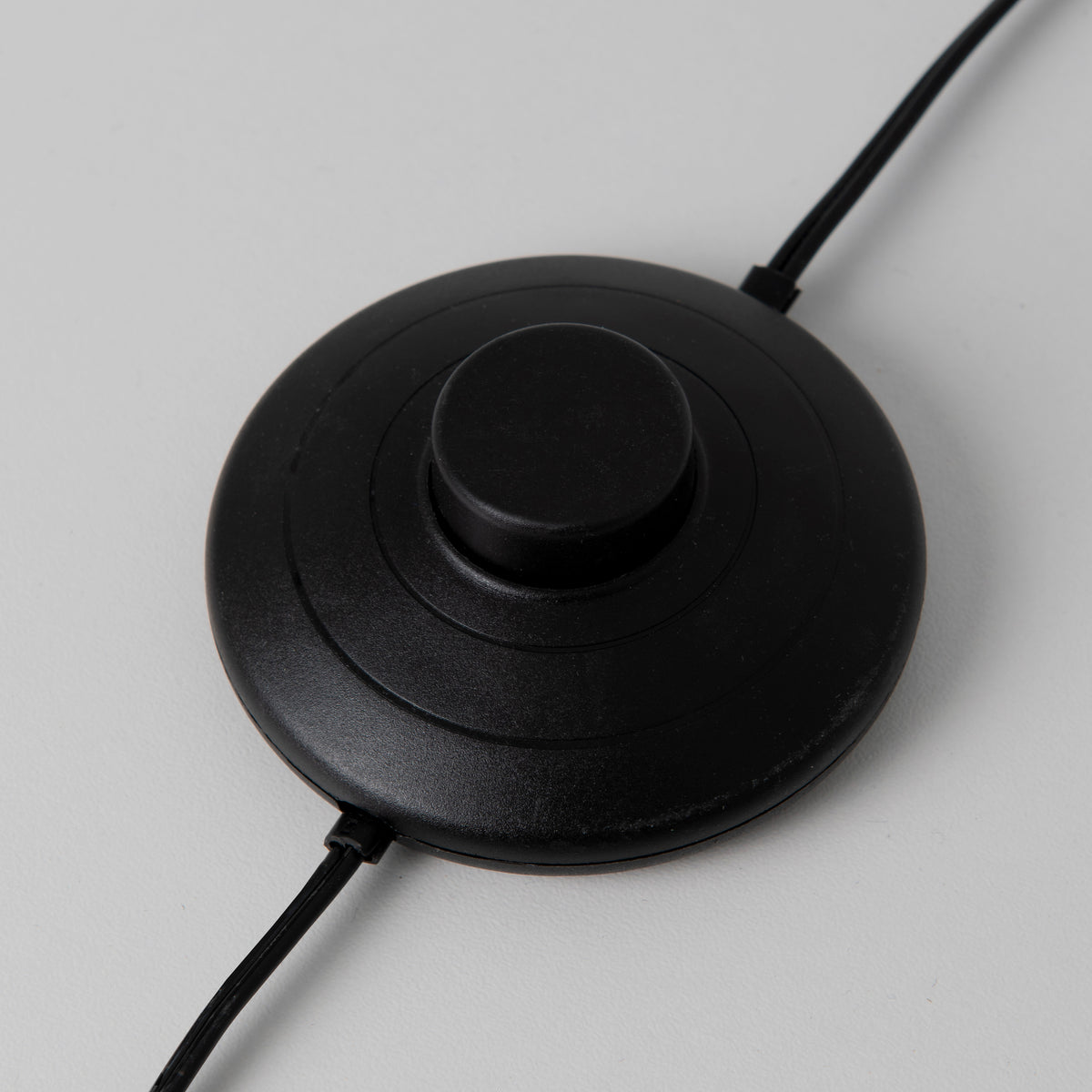 A round, black foot switch with a raised button in the center and two black cords extending from opposite sides, set against a light gray background.