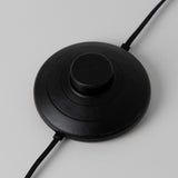 A round, black foot switch with a raised button in the center and two black cords extending from opposite sides, set against a light gray background.