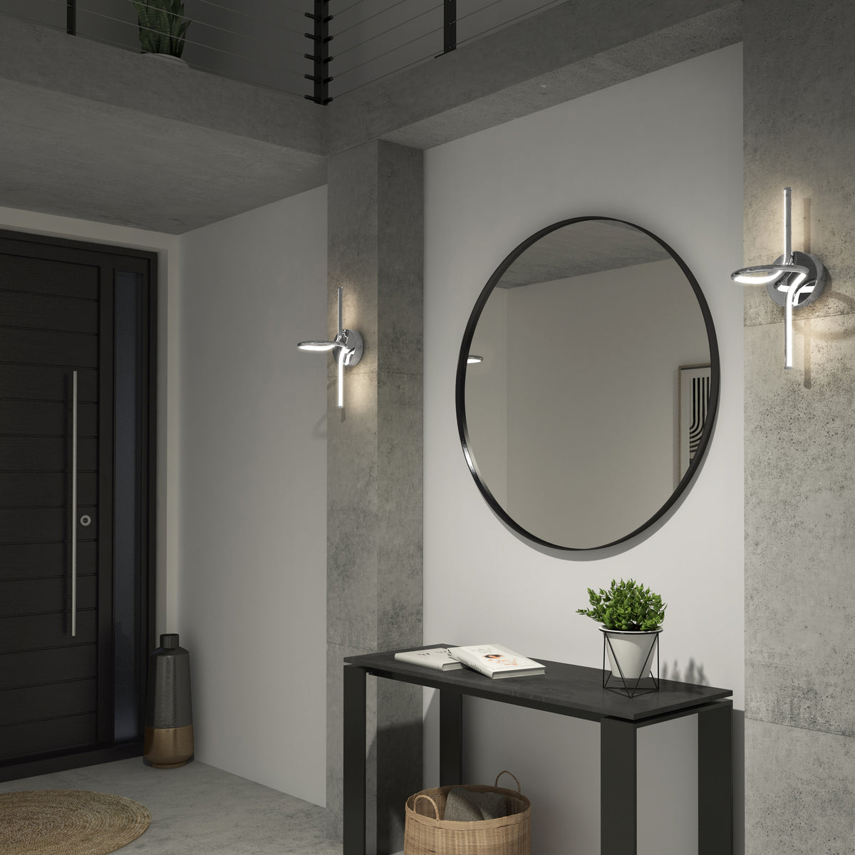 A modern entryway with concrete walls features a black console table, a round wall mirror, a potted plant, books, a basket underneath, two wall sconces, and a black front door in the background.