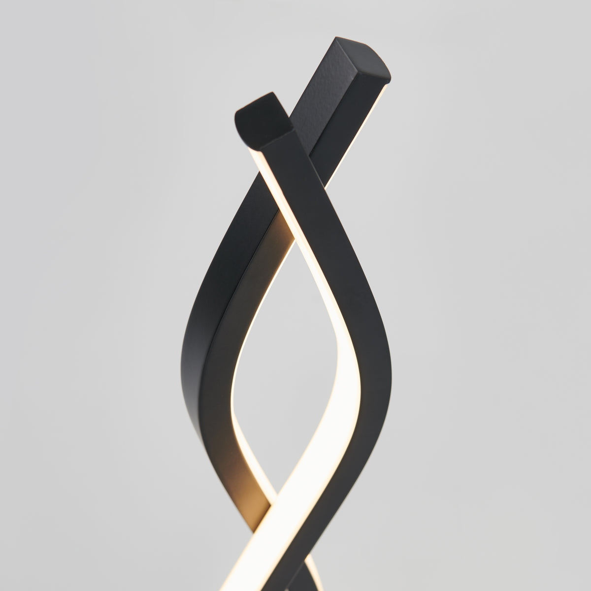 A modern table lamp with two twisting, black metal strips; one strip emits a soft white light, creating an elegant, sculptural design against a plain light gray background.