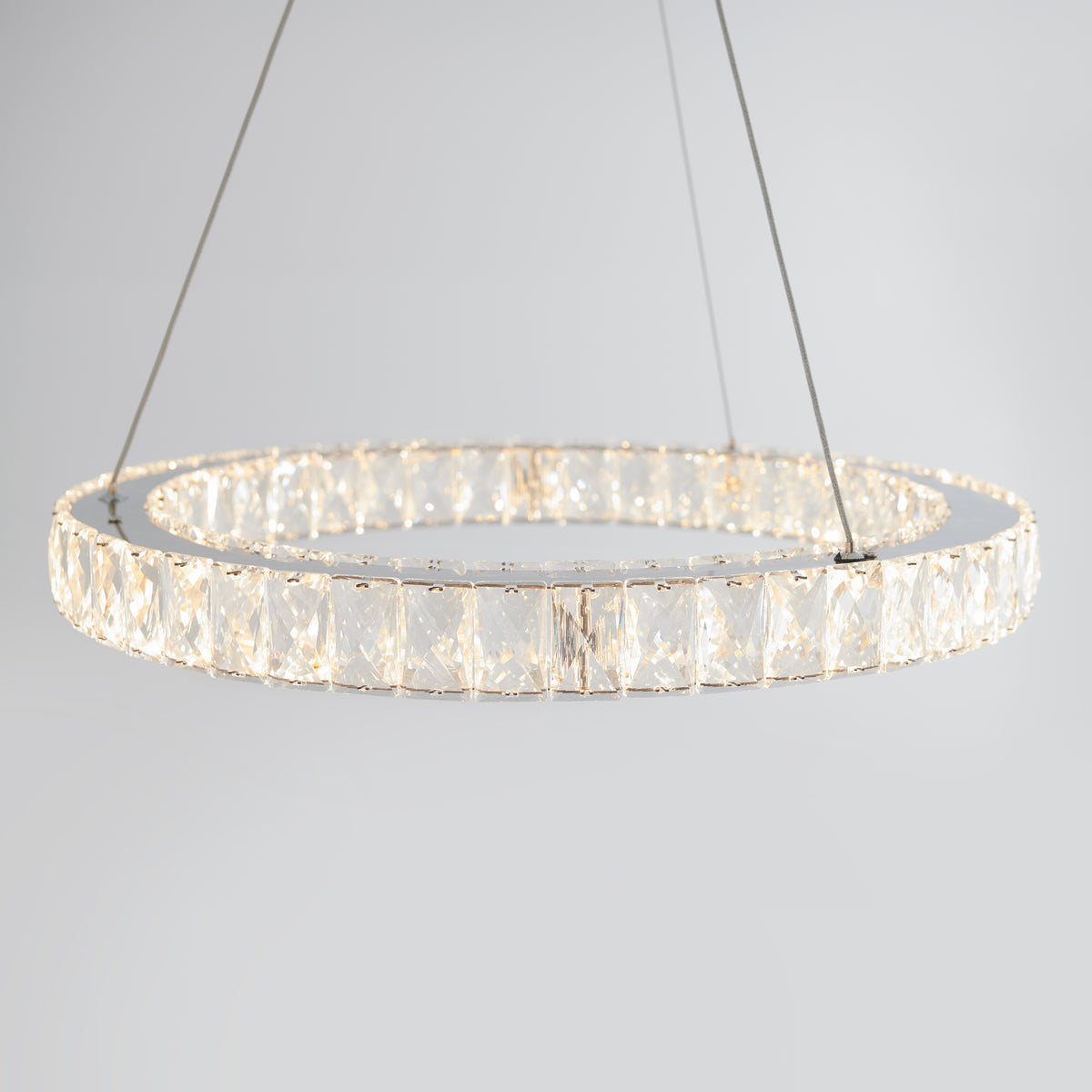 A modern circular chandelier with clear crystal accents, suspended by thin wires against a plain light gray background. The chandelier emits a soft, warm light.