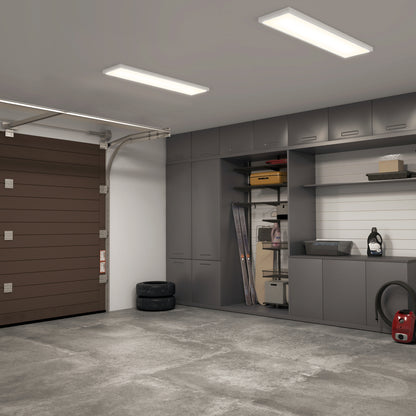 A clean, modern garage with gray cabinets and shelves is brightly lit by the Artika Skyshade smart RGB LED light panel. Tools and storage items are organized on shelves, with tires and a vacuum on the floor beside a brown garage door.