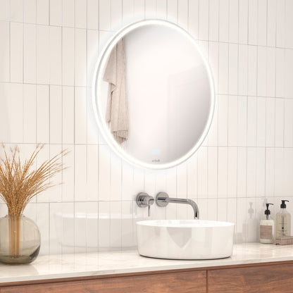 Modern bathroom featuring the Artika Emeraude anti fog LED wall mirror above a white sink with wall-mounted faucet, soap dispensers, and a glass vase with dried grass on a wooden countertop against light tile walls.
