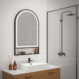 A modern bathroom with a wooden vanity, white sink, black faucet, and an arched LED mirror above. Glass shower enclosure with black fixtures is visible to the right. Brown bottles and a reed diffuser sit on the counter.