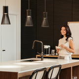 A woman stands in a modern kitchen, smiling and holding a bowl. The kitchen features a large island with a sink, stools, and pendant lights, with a dark textured wall in the background.