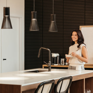 A woman smiles in a modern kitchen with black textured walls, a large island with sink and two chairs, illuminated by Artikas Kiyo modern LED pendant 3 CCT lights.