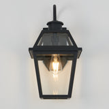 A black outdoor wall lantern with a clear glass enclosure and a visible light bulb, mounted on a white wall. The lantern has a modern, geometric design.