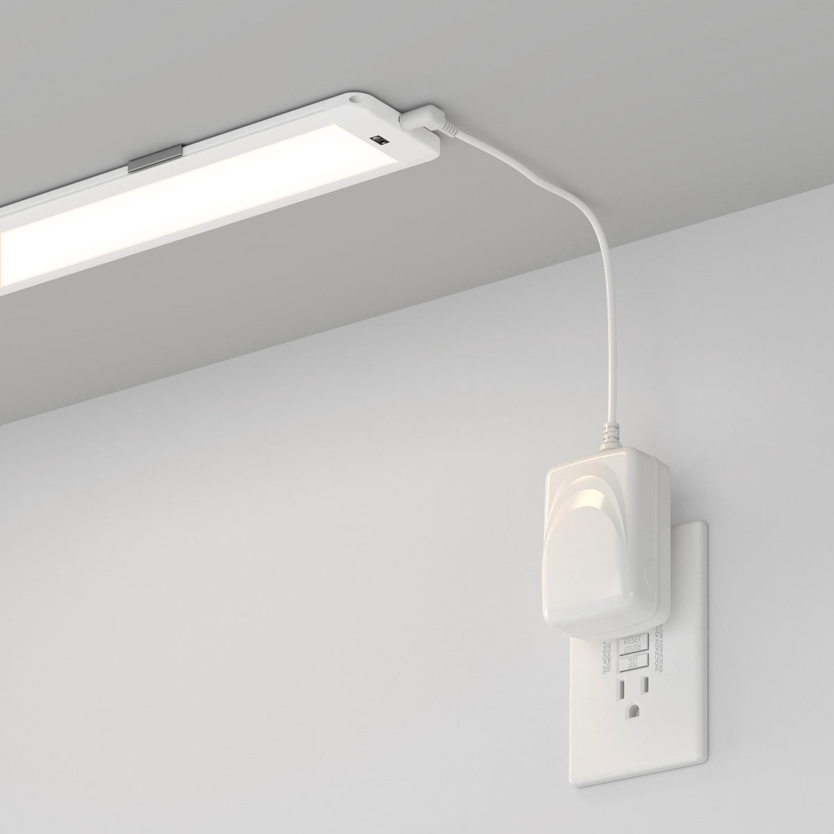 A rectangular LED light fixture is mounted under a shelf or cabinet, plugged into a white electrical outlet on the wall using a visible white power adapter and cord.