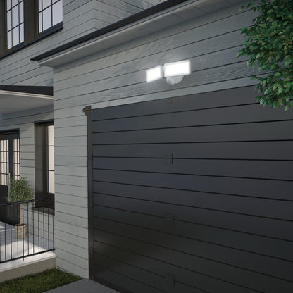 A modern garage with dark horizontal panels is attached to a light gray house. Two Artika Sentinel outdoor Flat Panel Security lights are mounted above the garage door beside a leafy green tree.