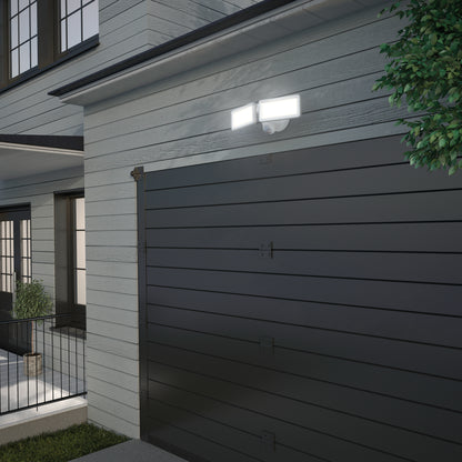 The Artika Sentinel outdoor Flat Panel Security light is mounted above a dark garage door on a modern house, lighting up the exterior siding in the evening. A small tree branch appears in the upper right corner.