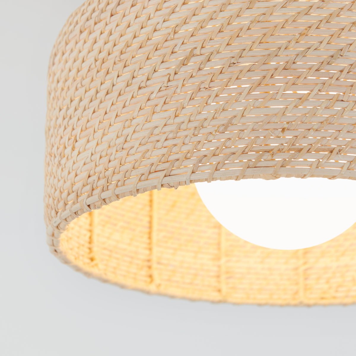 Close-up of a woven rattan lampshade with a light bulb glowing inside, against a plain light background. The texture of the natural fibers is clearly visible.