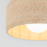 Close-up of a woven rattan lampshade with a light bulb glowing inside, against a plain light background. The texture of the natural fibers is clearly visible.