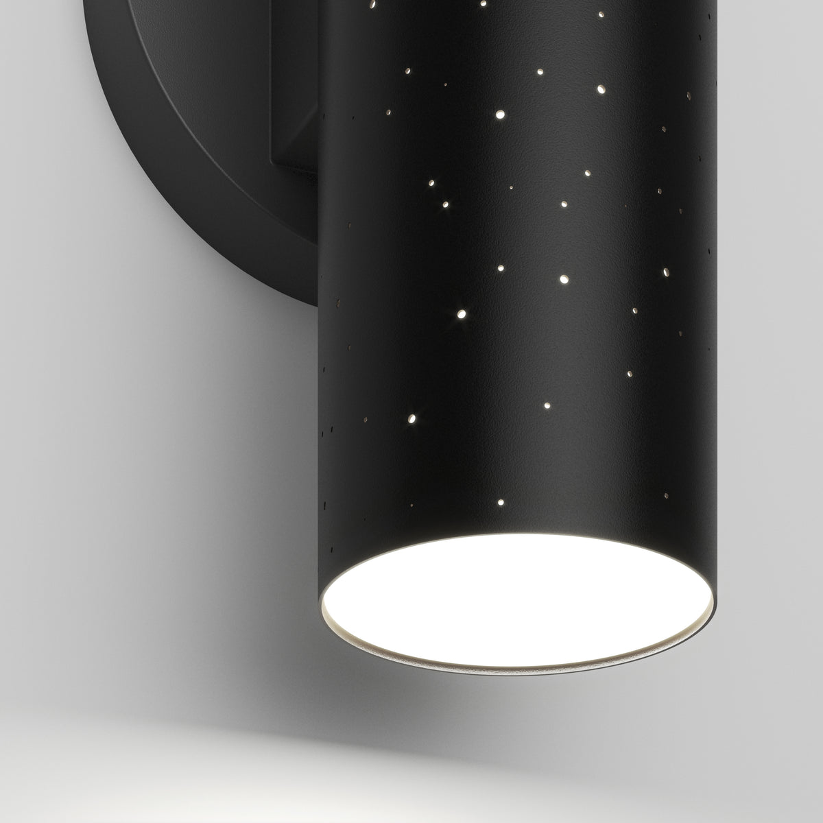 A close-up of Artika’s Cosmic indoor-outdoor LED wall light 3 CCT, a black cylindrical fixture with star-like perforations, emitting light downward and mounted on a white wall.