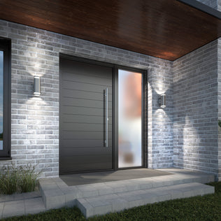 Modern front entrance with a dark horizontal panel door, large vertical frosted glass side panel, gray brick walls, Artika’s Mettle LED indoor-outdoor wall light, and concrete steps edged with grass.