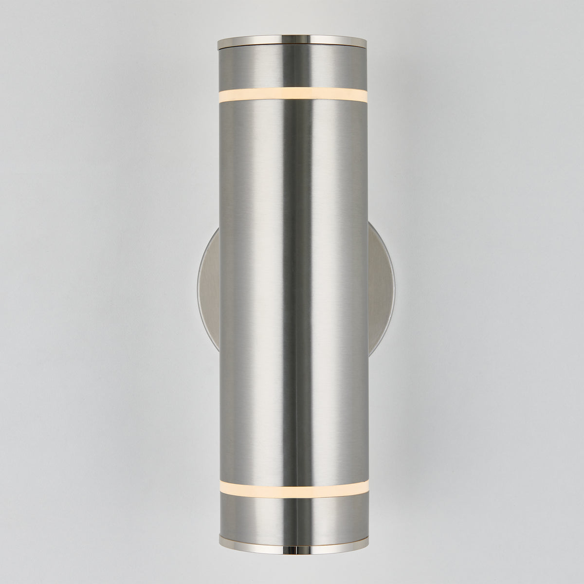 The Artika C7 indoor-outdoor wall light is a modern cylindrical brushed metal sconce, emitting soft white light from narrow top and bottom bands, mounted on a circular base against a plain light background.