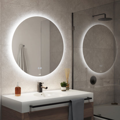 A modern bathroom featuring the Artika Raya 24 in. LED mirror with defogger above the sink, a towel on the wall, glass shower enclosure, and minimalistic decor such as a plant and soap dispenser on the countertop.