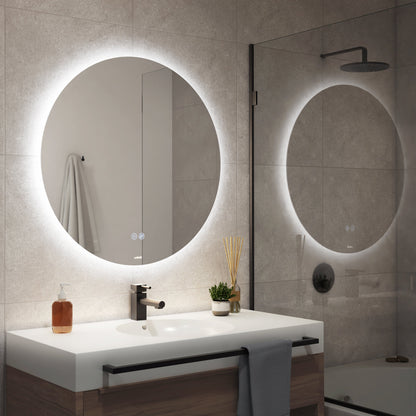 A modern bathroom features the Artika-CAN-dev Raya 24 in. LED mirror (3 CCT, defogger) above a white sink with a black faucet, counter towel, soap dispenser, plant, reed diffuser; wall towel and shower also visible.